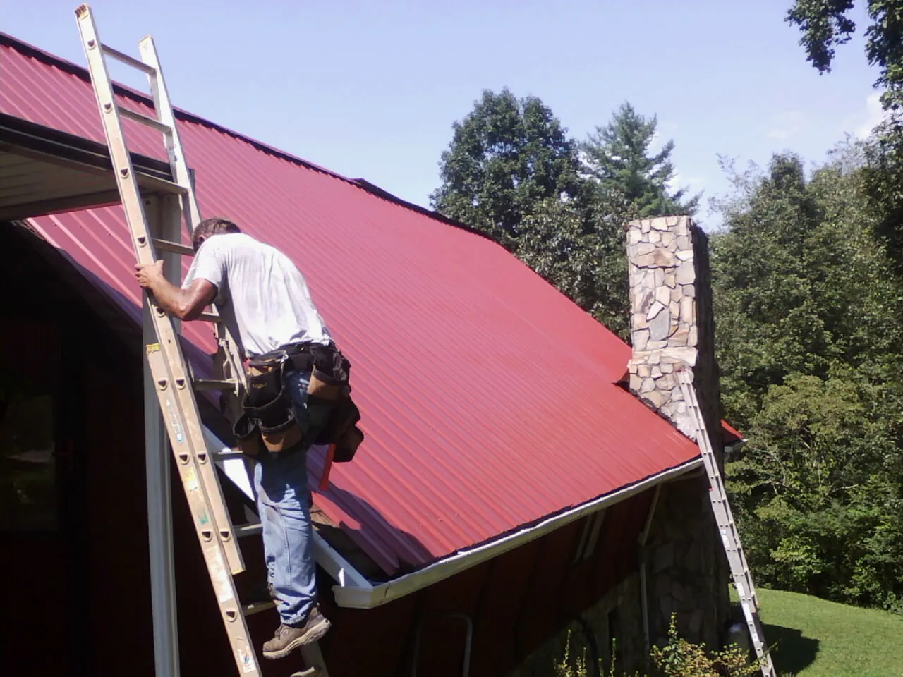 Roofer on a ladder beside a red metal roof and stone chimney — Soffit & Fascia Repair by Roofing Brothers in Cincinnati